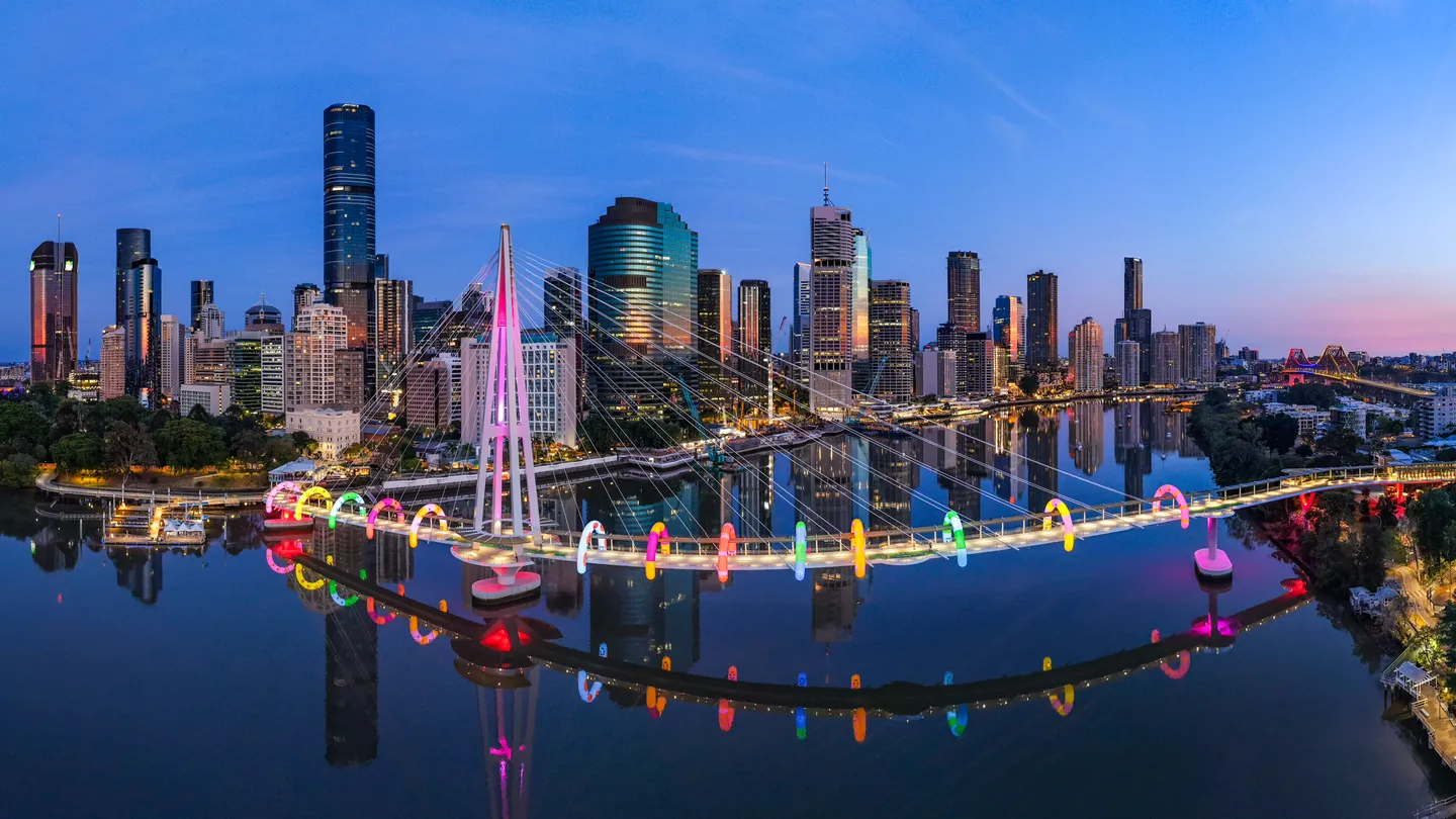 An aerial view of Brisbane city at dawn with Kangaroo Point Bridge lit up with rainbow arches of ANZ's Walk This Way by Craig Karl. 