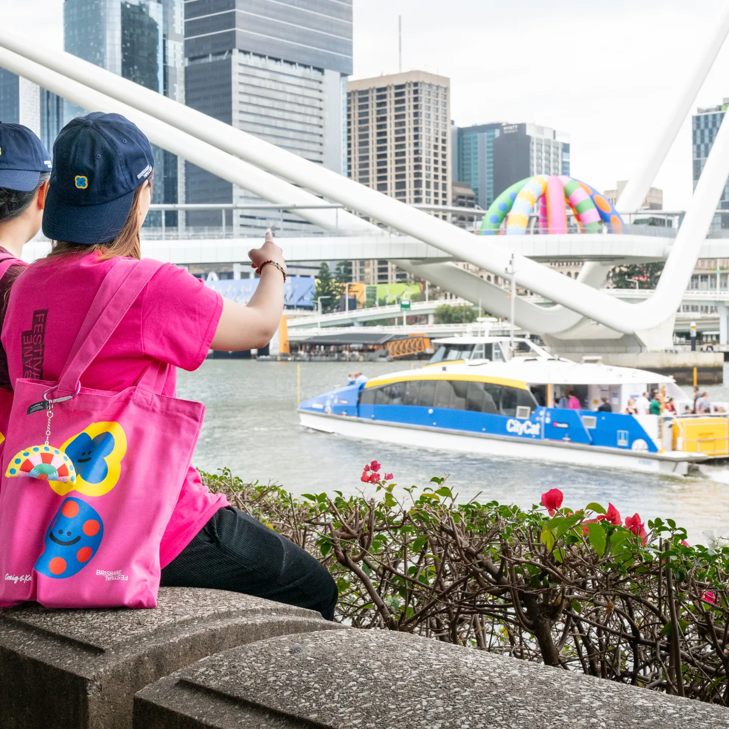 Two people with Criag & Karl merchandise point at a Citycat Ferry and at an art installation on a bridge