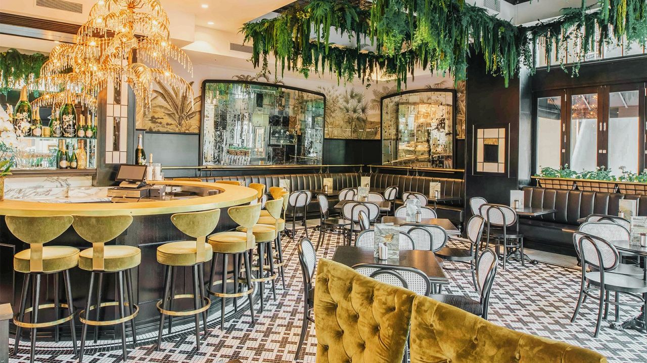 An art-deco inspired bar and dining space features gold plush chairs, a gold fountain light installation, a circle bar and green plants draping down from the ceiling. 