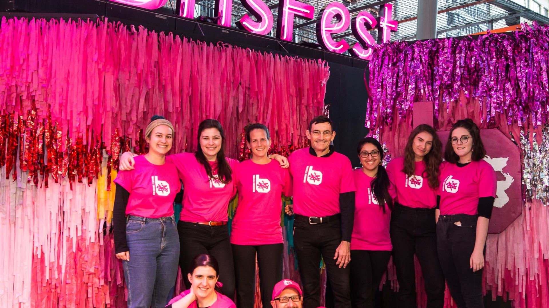 A group of people in pink Brisbane Festival shirts in front of a large pink tassel backdrop and "#BrisFest" light installation.